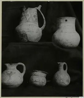 Five pottery vessels, including jugs, a jar and a bowl, arranged on dark draped cloth in two tiers