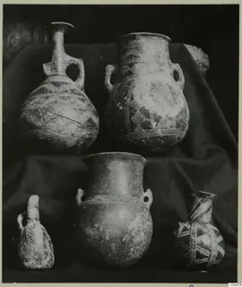 Five pottery vessels, jugs and jars with painted and relief decoration, grouped on dark draped cloth