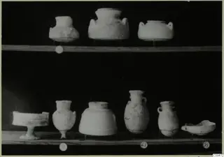 Several small pottery vessels and bowls standing on two wooden shelves against a black background.