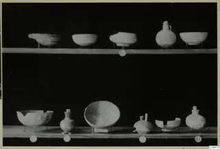 Eleven pale vessels, bowls and small jugs, set out on two wooden shelves against a black background