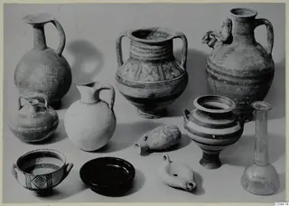 Group of about a dozen pottery vessels, a glass bottle and an oil lamp on a pale surface