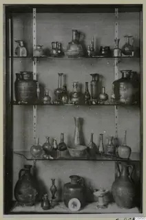 Glass vessels of many shapes displayed on four shelves in a glass-fronted case.