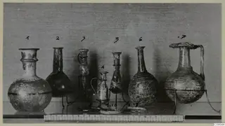 Seven blown glass vessels in a row on wire stands, numbered, with a scale bar along the front of the shelf.