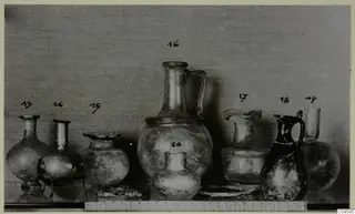 Group of eight blown glass jugs, jars and vases with numbered labels, photographed against a grey backdrop.
