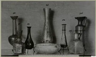 Row of blown glass jugs, vases and a ribbed bowl, each vessel with a handwritten number label above or beside it.