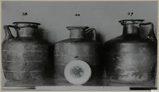 Three jugs with handles photographed in a row, with number labels and a white round dish in front