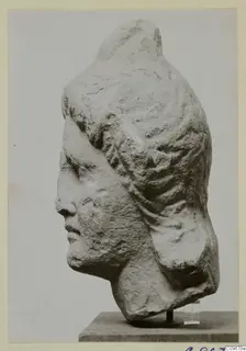 Carved stone head of a figure with hair swept back in ridged waves, shown in profile on a stand.