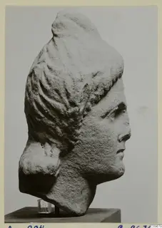 Stone head of a figure in profile facing right, with hair drawn back in wavy strands, on a dark base
