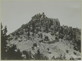 Rocky hilltop with ruined walls and towers running down its slopes, with pines in the foreground.