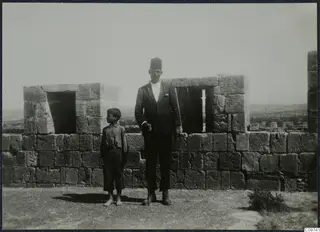 A man in a suit and a tall flat-topped cap with a cane, and a barefoot boy, standing on a stone fort wall at Kolossi