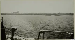 Buildings along a long sea wall, seen across calm water from the rail of a ship