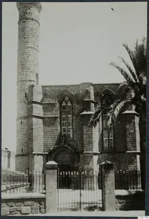 Stone building with pointed-arched stained glass windows, a tall round tower and an iron gate, Nicosia.