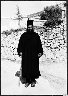 Papa Polivios, a bearded man in a long black robe and tall black hat, standing with a walking stick in a village street by a stone wall.