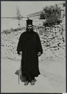 Papa Polivios standing on a dirt track in Mandres, Ayios Iakovos, holding a walking stick, with a dry-stone wall behind him.