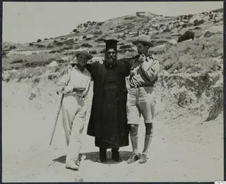 Three men stand arm in arm on a dirt track in Mandres, one a bearded priest in a long black robe and tall black hat.
