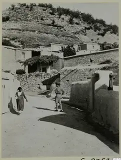 Village street at Lindros with mud-brick and stone walls, a woman walking past and a man in shorts standing in the road.
