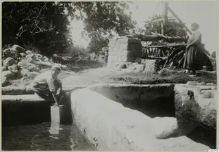 A man crouched at a stone-lined water channel, dipping a tin into the water, with a woman standing by a wooden wheel frame.