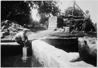 Man crouching to dip a container into a stone water channel, with a woman standing beside a wooden wheel frame and a stone structure.