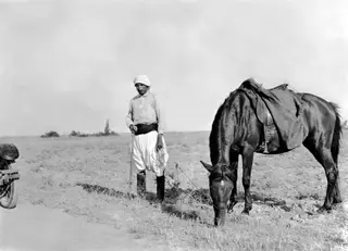 A man in a light head cloth and loose white trousers holds the rein of a grazing dark horse on open flat ground.
