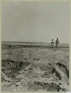 Two people stand on a mound above excavated stone foundations at Kalopsida, with flat open land behind them.