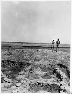 Two people stand on a mound beside stone house foundations at Kalopsida, with flat plain and hills behind.