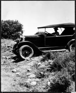 Open-topped car standing on a stony slope, with a driver at the wheel and bushes beside it