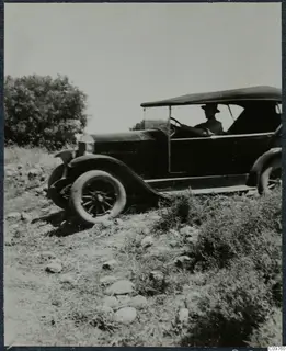 Open-topped vintage car with a driver on a stony slope beside bushes