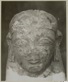 Frontal photograph of a weathered limestone sculpted head with side locks, against a dark background.