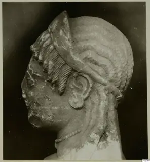 Carved stone head in profile from Arsos, with elaborate hair and a bead necklace.
