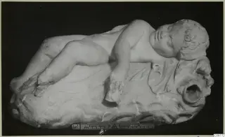 Marble sculpture of a sleeping child lying on a rocky base, with a ruler placed beside it for scale.