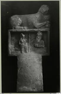 Carved stone object with a recumbent animal on top and a relief panel showing two seated figures
