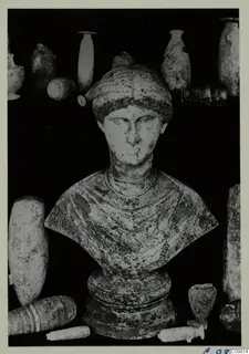 Roman sculpture: a bust of a figure with a head covering, set among several vessels on a dark background