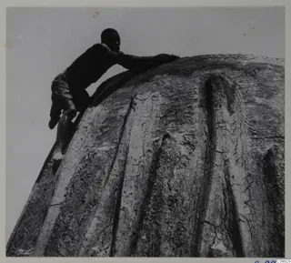 Man leaning over the curved top of a domed building with external ribs, photographed from below against the sky
