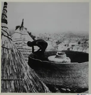 Man building a Matakam hut, with thatched conical roofs and open country with trees beyond.