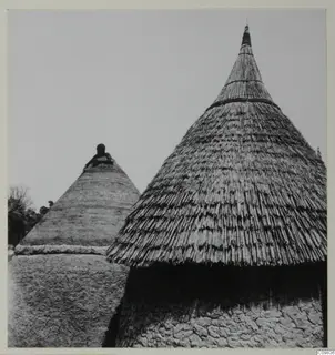Two conical thatched huts with mud walls, photographed from below against an open sky