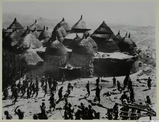 Conical thatched huts inside a walled settlement at Duvangar, with a crowd of people on bare ground in front.