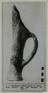 Pottery vessel with a tall pointed neck, a single loop handle and a wide flat base, shown in a photo mounted on a printed page