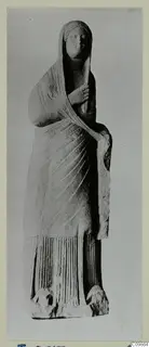 Standing draped female statue in stone, veiled, one hand raised to the chest.