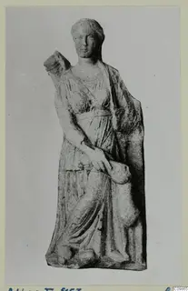 Standing draped female statue with her hair bound up, one arm raised to a block at her shoulder, shown on a grey card mount