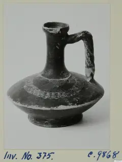 Dark jug with a tall neck, single strap handle and flaking pale paint bands on the body