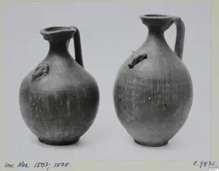 Two dark pottery jugs with single handles and applied knobs on the body, standing side by side