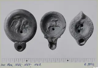 Three terracotta oil lamps with moulded decoration on the discs, shown side by side on a grey card with a scale below.