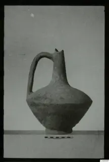 Pottery jug with a strap handle, narrow neck and broad body, photographed on a ledge with a scale bar