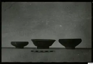 Three dark pottery bowls of different sizes with short feet, standing in a row with a scale bar in front.