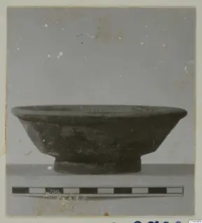 Side view of a dark pottery bowl with a flared rim and short ring foot, with a scale bar in front.