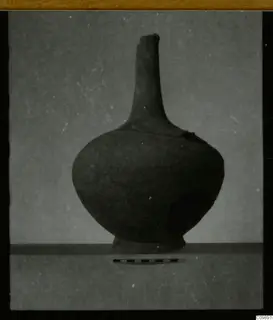 Pottery jar with a tall narrow neck and rounded body, photographed on a shelf beside a scale bar
