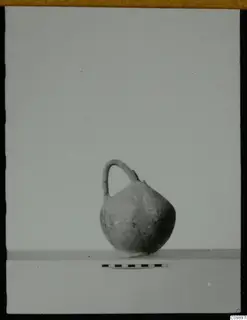 Small round-bodied jug with a single loop handle, standing on a shelf beside a scale bar.
