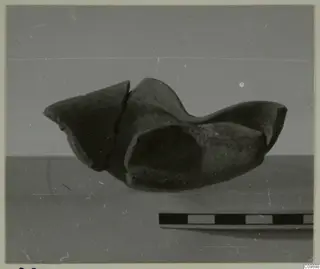 Fragmentary dark pottery vessel with broken rims, shown beside a scale bar against a grey backdrop.