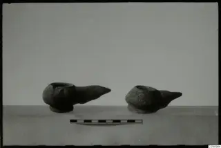 Two dark oil lamps with pointed nozzles standing on a pale surface, with a scale bar in front.