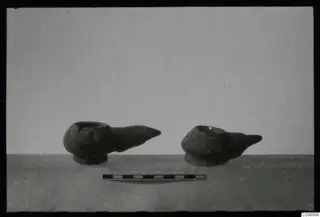 Two oil lamps with pointed spouts standing on a surface, with a scale bar in front